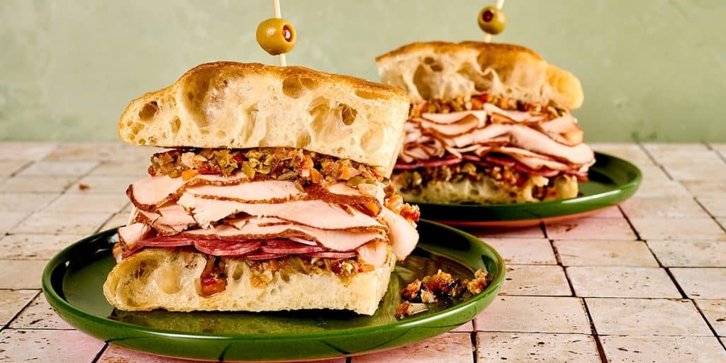 A platter of sliced muffuletta deli sandwiches layered with cured meats, cheeses, and olive spread.